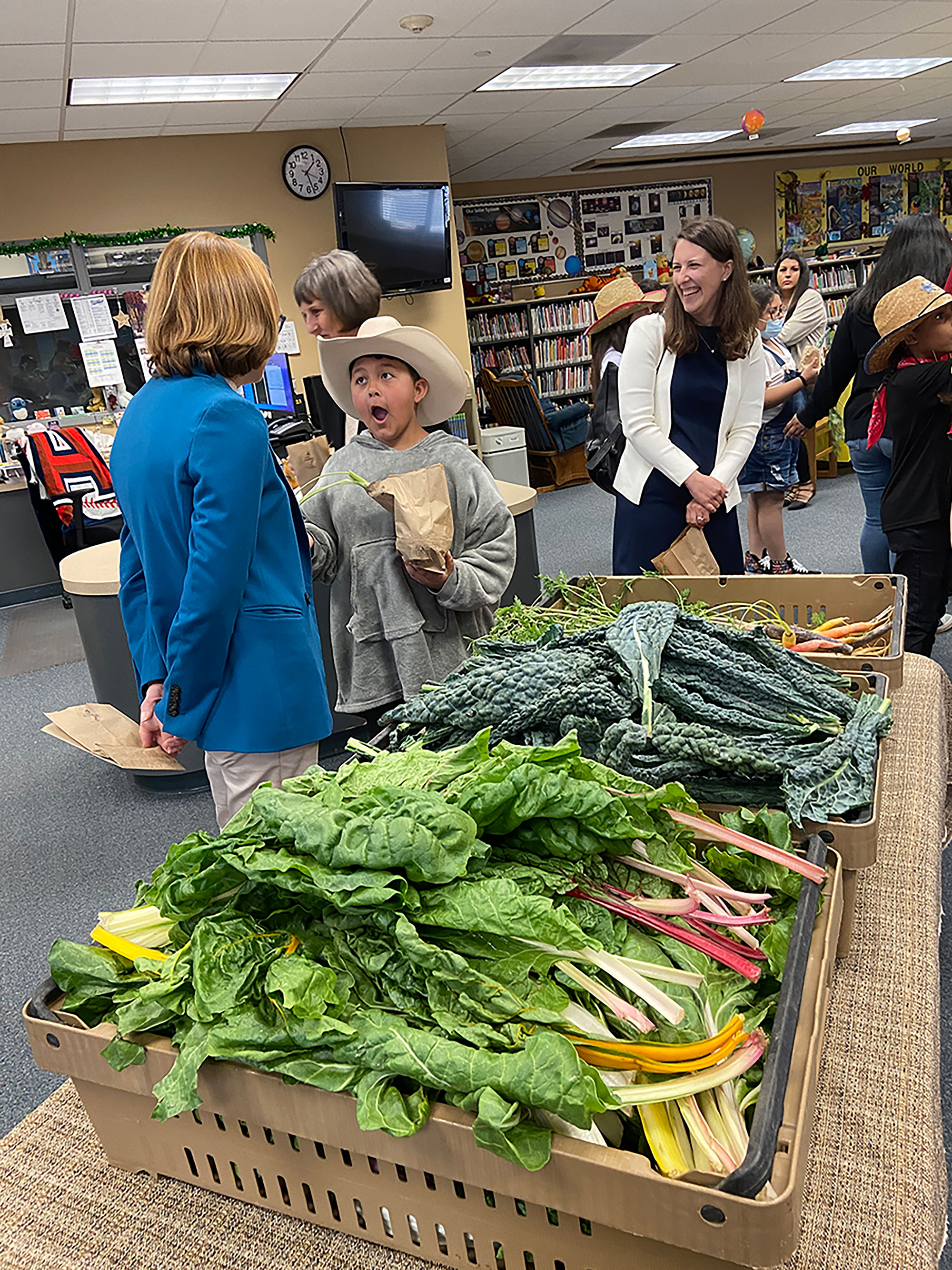 FNS Administrator Cindy Long speaks with a surprised Werner Elementary School student wearing a wide-brimmed hat standing next to boxes of vegetables inside a school library