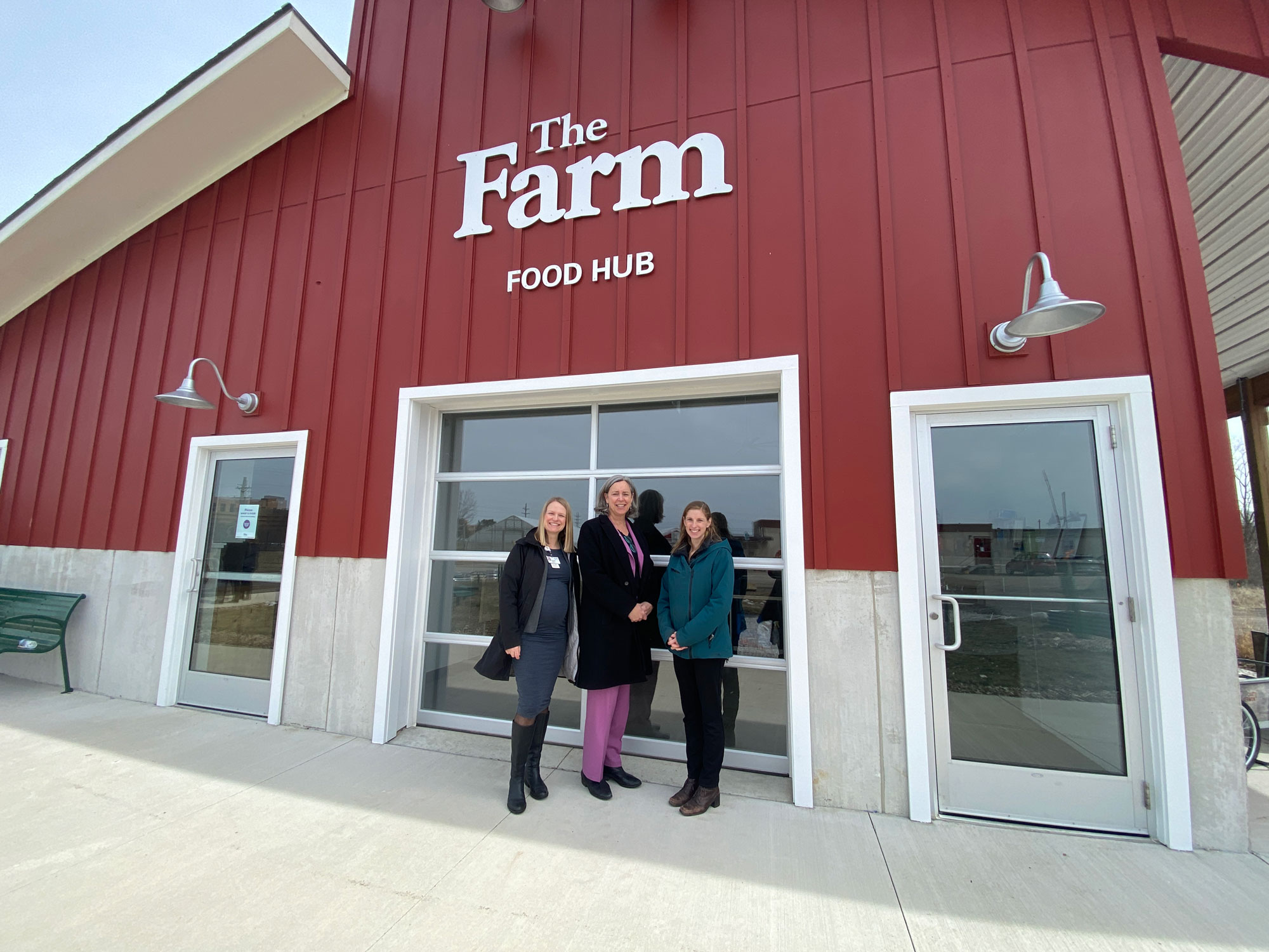 Amanda Sweetman, Stacy Dean, and Jae Gerhart stand in front of a building that has a sign on it that reads: The Farm – Food Hub