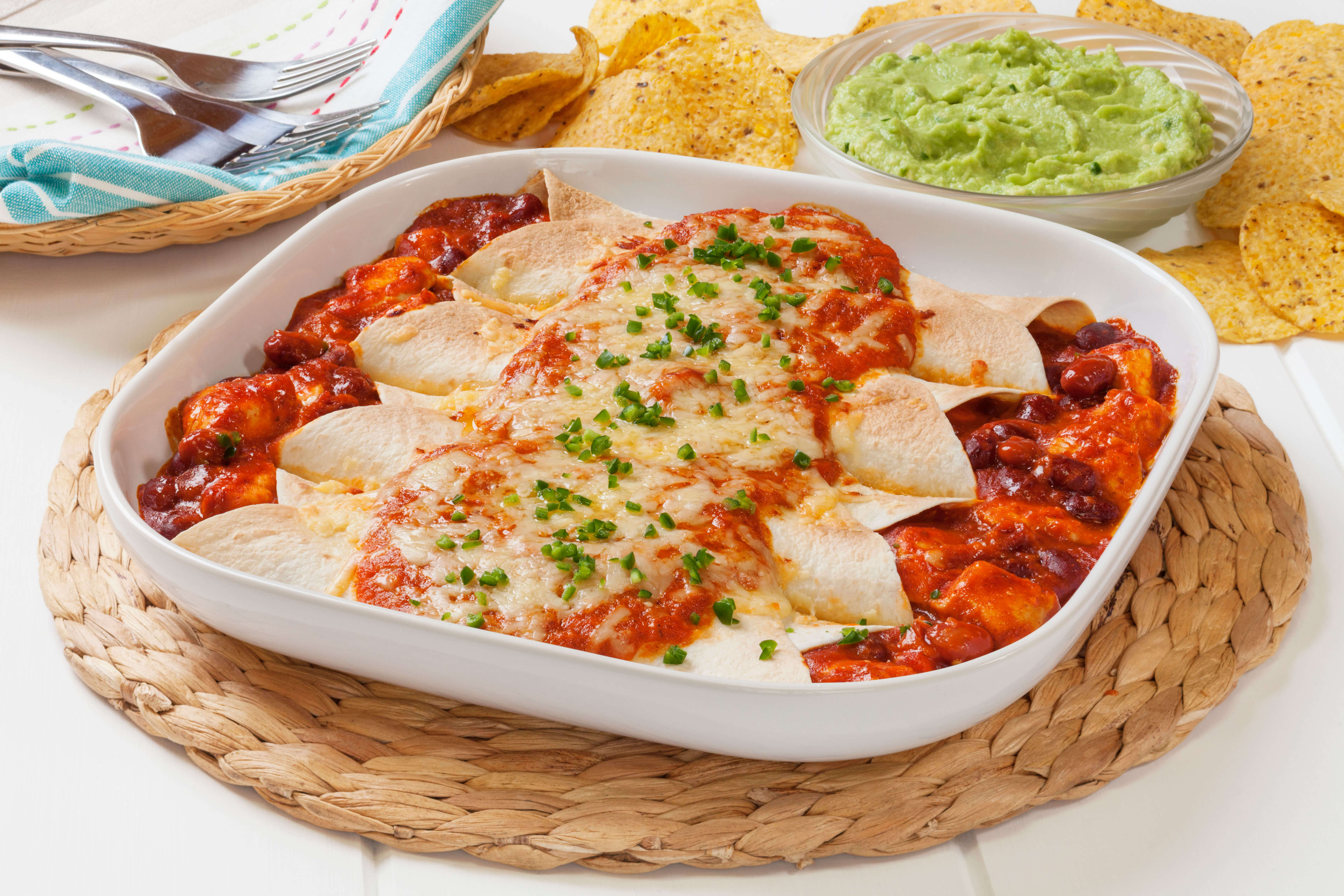 A white serving platter filled with enchiladas rests on a woven trivet. Round tortilla chips, a bowl of guacamole, and utensils are near the platter