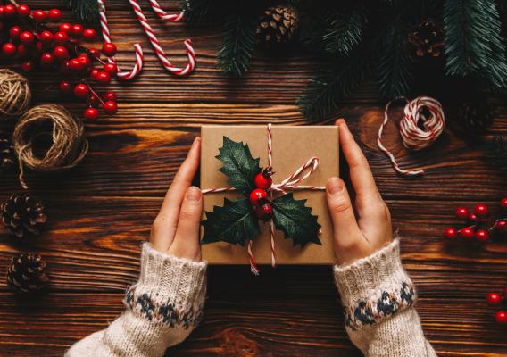 Adobe Stock image by Pink Coffee studio depicts a person holding a holiday package wrapped with holly. Other decorations are neatly stacked around the package, including pine branches, cones, twine, berries and sugar canes