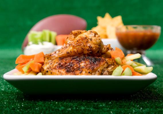 Close up of a full plate of spicy chicken wings with celery and carrot sticks along with championship game party items in the background