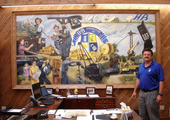 Robert Koch, President and General Manager of H&B Communications in Holyrood, Kansas, stands in front of a mural highlighting milestones in his family's telecommunications business.